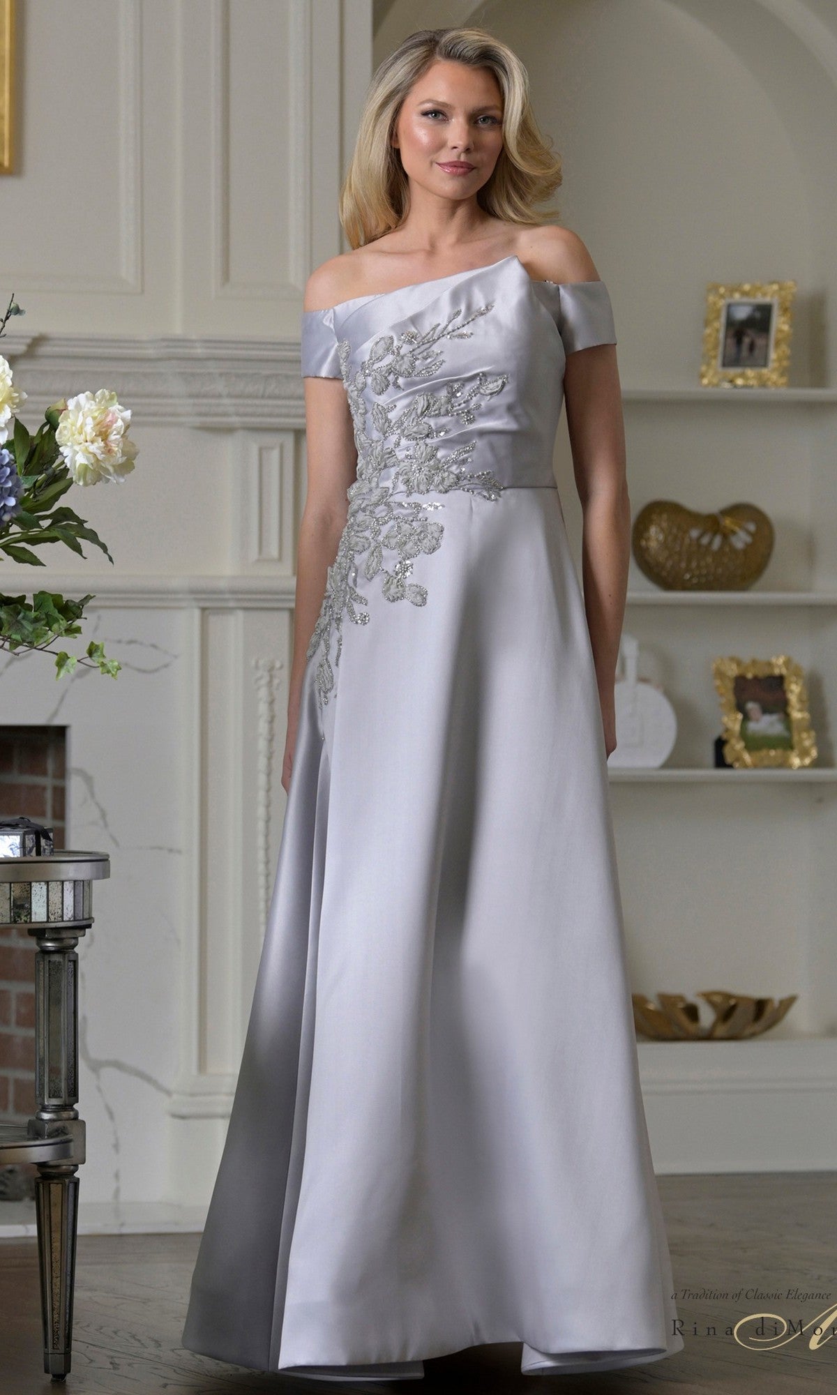 A woman in the silver Rina Di Montella RD3501 long formal dress, featuring an A-line skirt and crafted from shimmering Mikado with floral embroidery, stands indoors by a fireplace and decorative shelves.